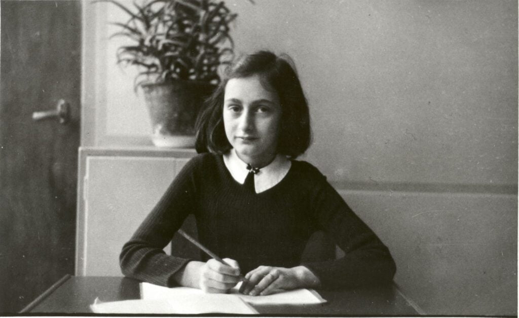 Na foto: Anne Frank sentada escrevendo em um papel. Imagem cortesia da "Anne Frank Fonds"