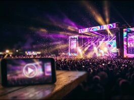 Palco de show iluminado com luzes roxas e celular exibindo transmissão ao vivo